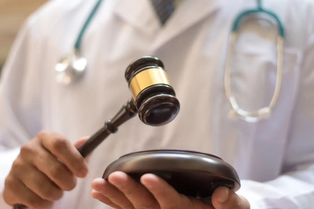 A person in a white coat and stethoscope holds a gavel poised over a sound block symbolizing the intersection of law and medicine. The focus is on the hands and the gavel with the rest of the figure blurred.